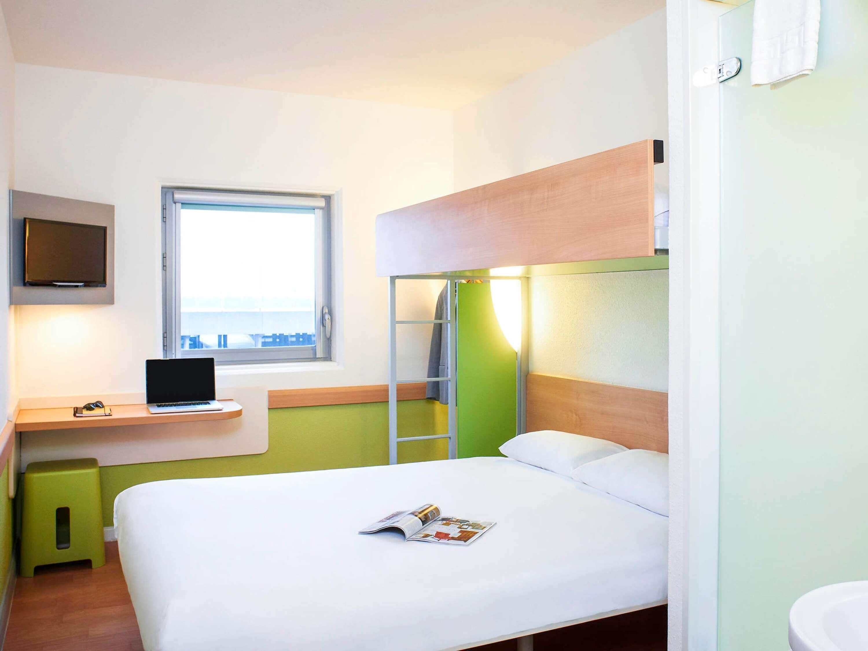 A hotel room with a bed, a desk with a laptop, a TV mounted on the wall, and a window with a view.