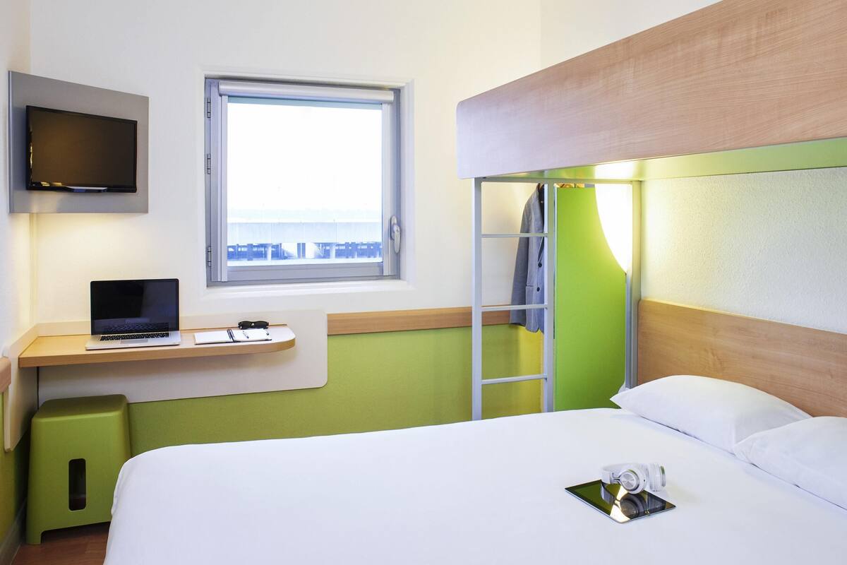 A hotel room with a bed, a desk with a laptop, a TV, and a window with a view.