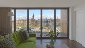 Ein modernes Wohnzimmer mit einem grünen Sofa, einem Couchtisch mit einer Vase und Blick auf eine Stadtkulisse durch große Fenster.