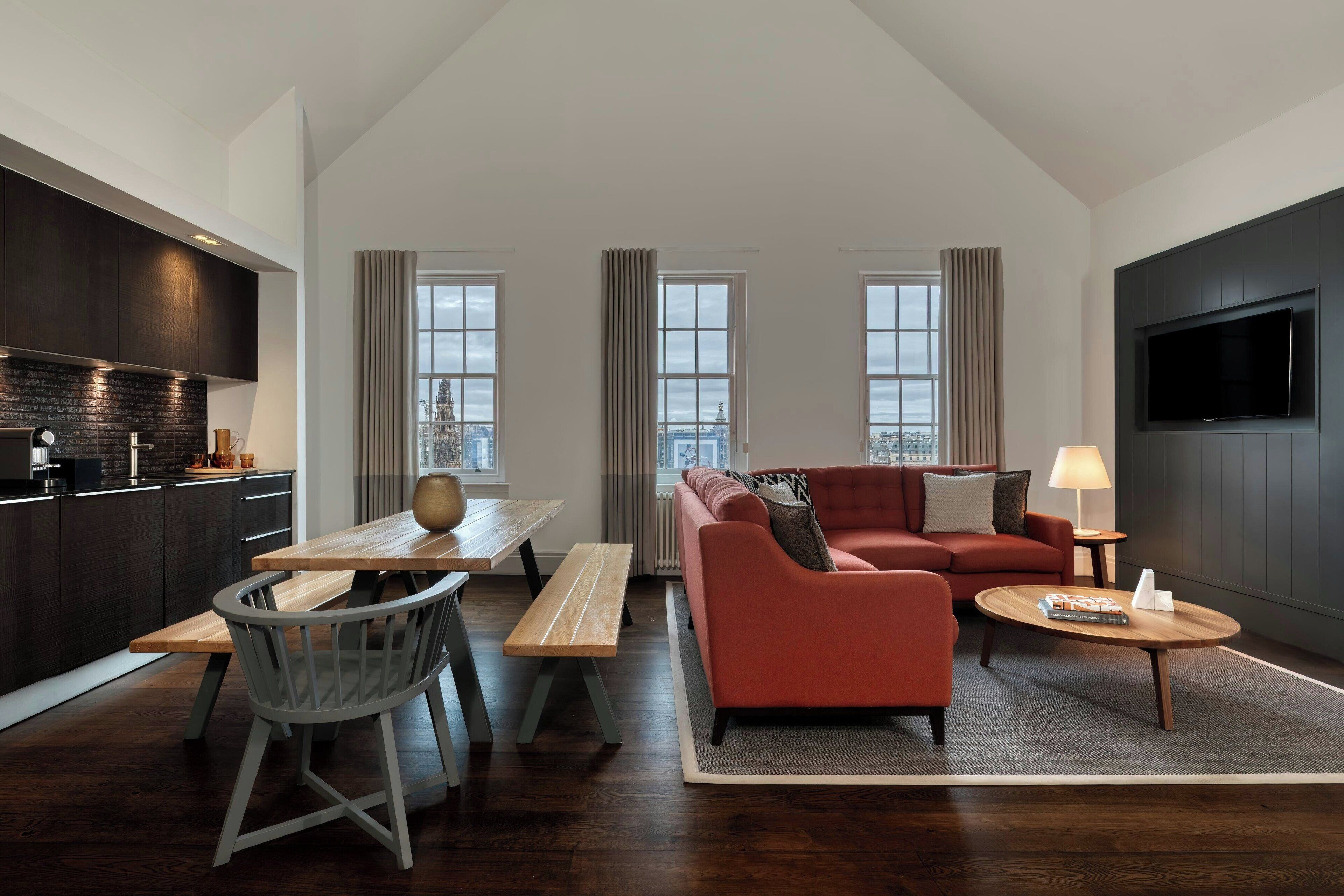 A modern living room with a sofa, a coffee table, a dining area with a bench and chairs, a kitchen with dark cabinets, and a large TV mounted on the wall.