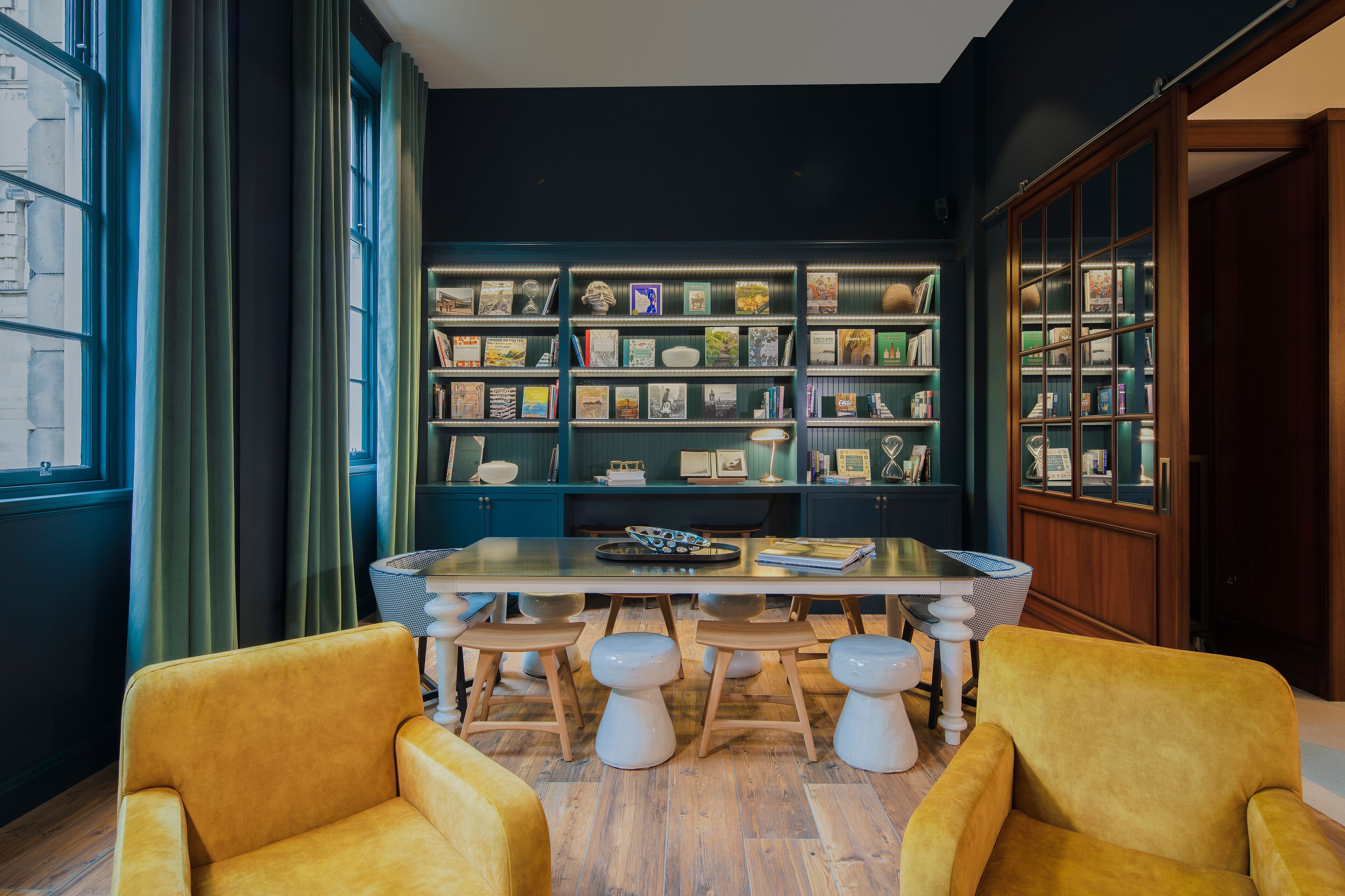 A room with a wooden table, four stools, a bookshelf, and a large window with curtains.