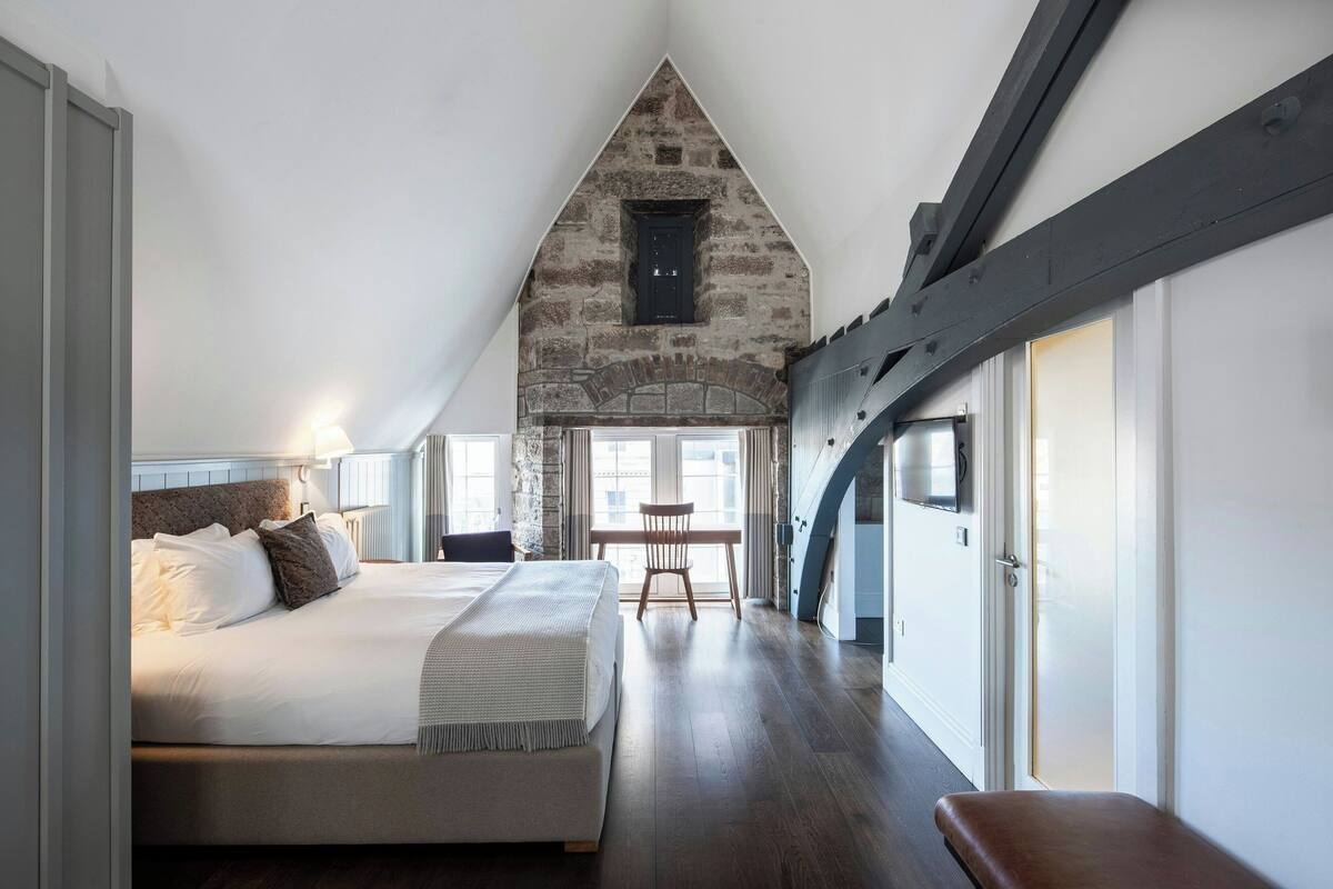 A bedroom with a large bed, a wooden bench, a desk, and a stone wall.