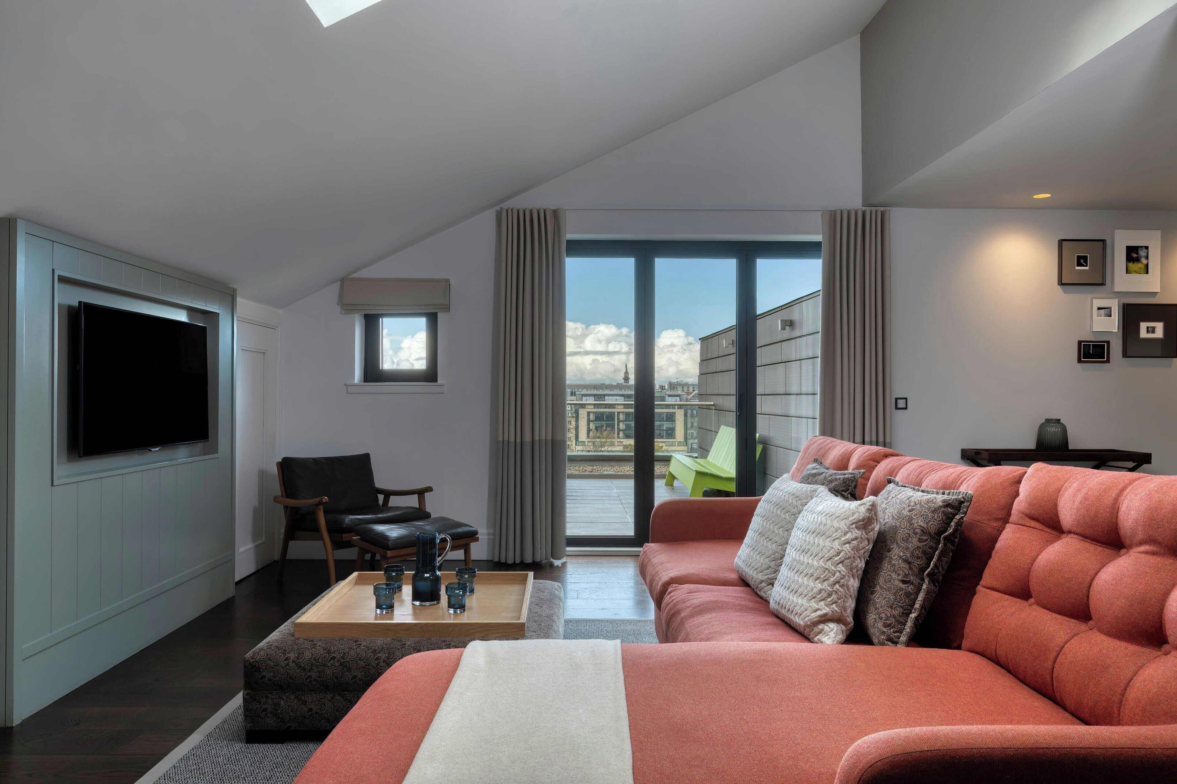A modern living room with a large flat-screen TV, a sofa, a coffee table, and a view of a cityscape through large windows.