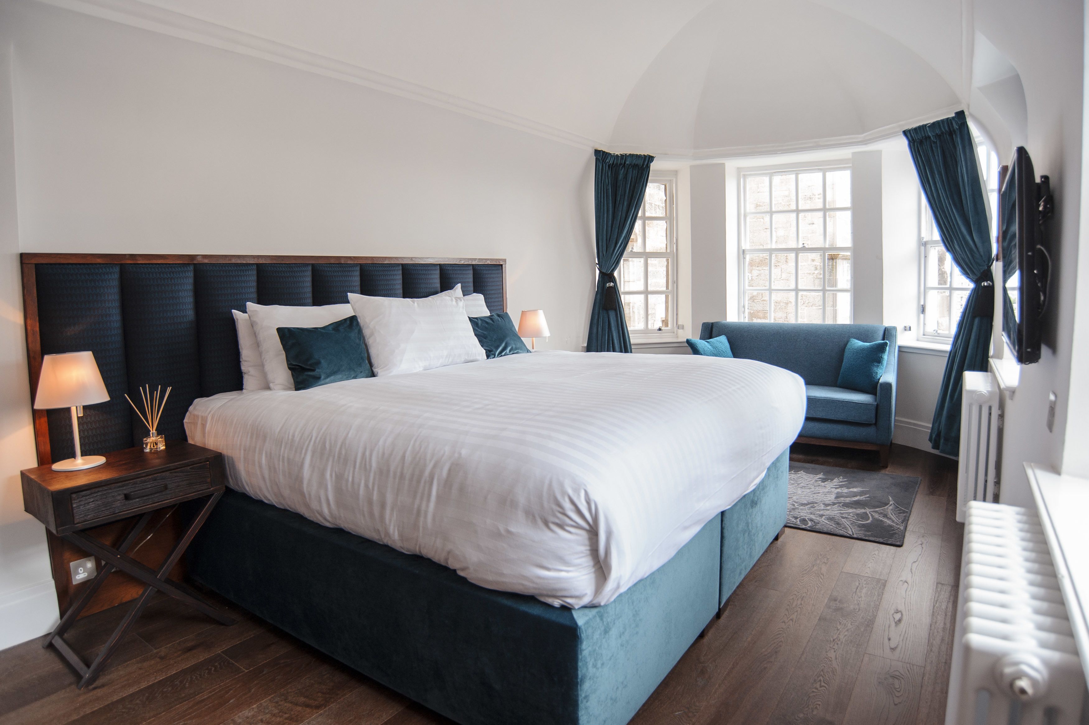A large bed with a dark headboard, a bedside table with a lamp, and a blue armchair.