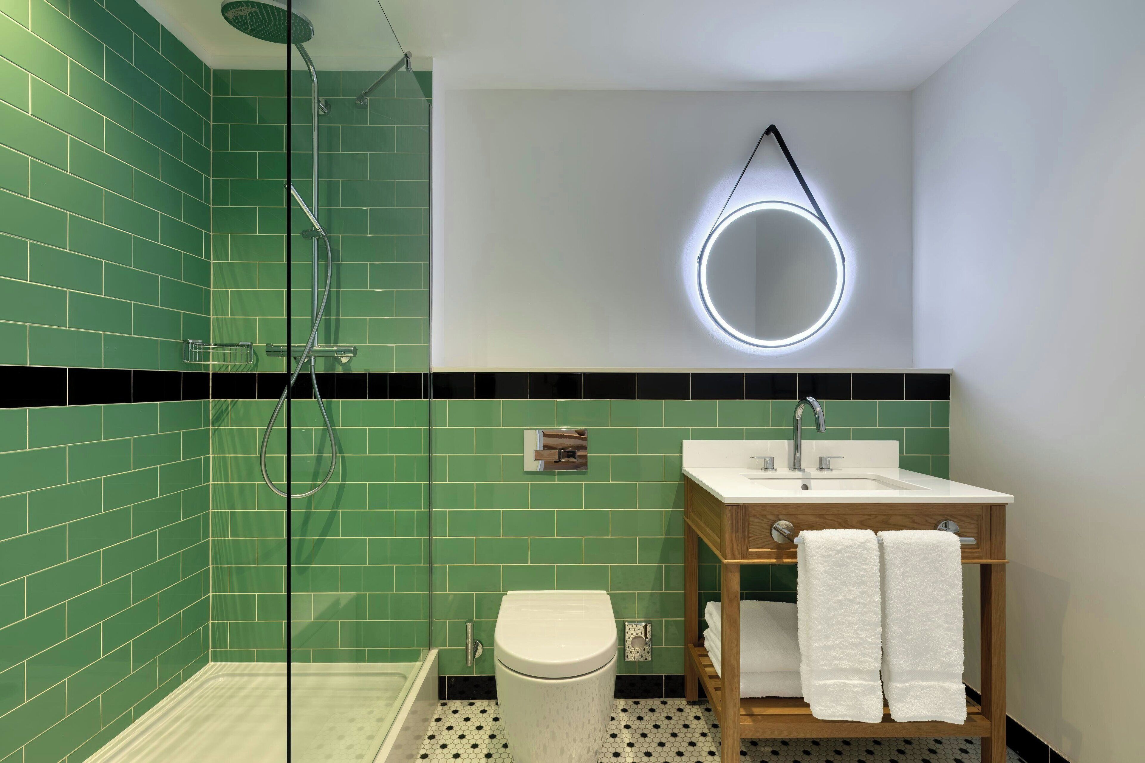 A bathroom with green tiles, a glass shower enclosure, a toilet, a sink with a wooden vanity, and a round mirror with a light.