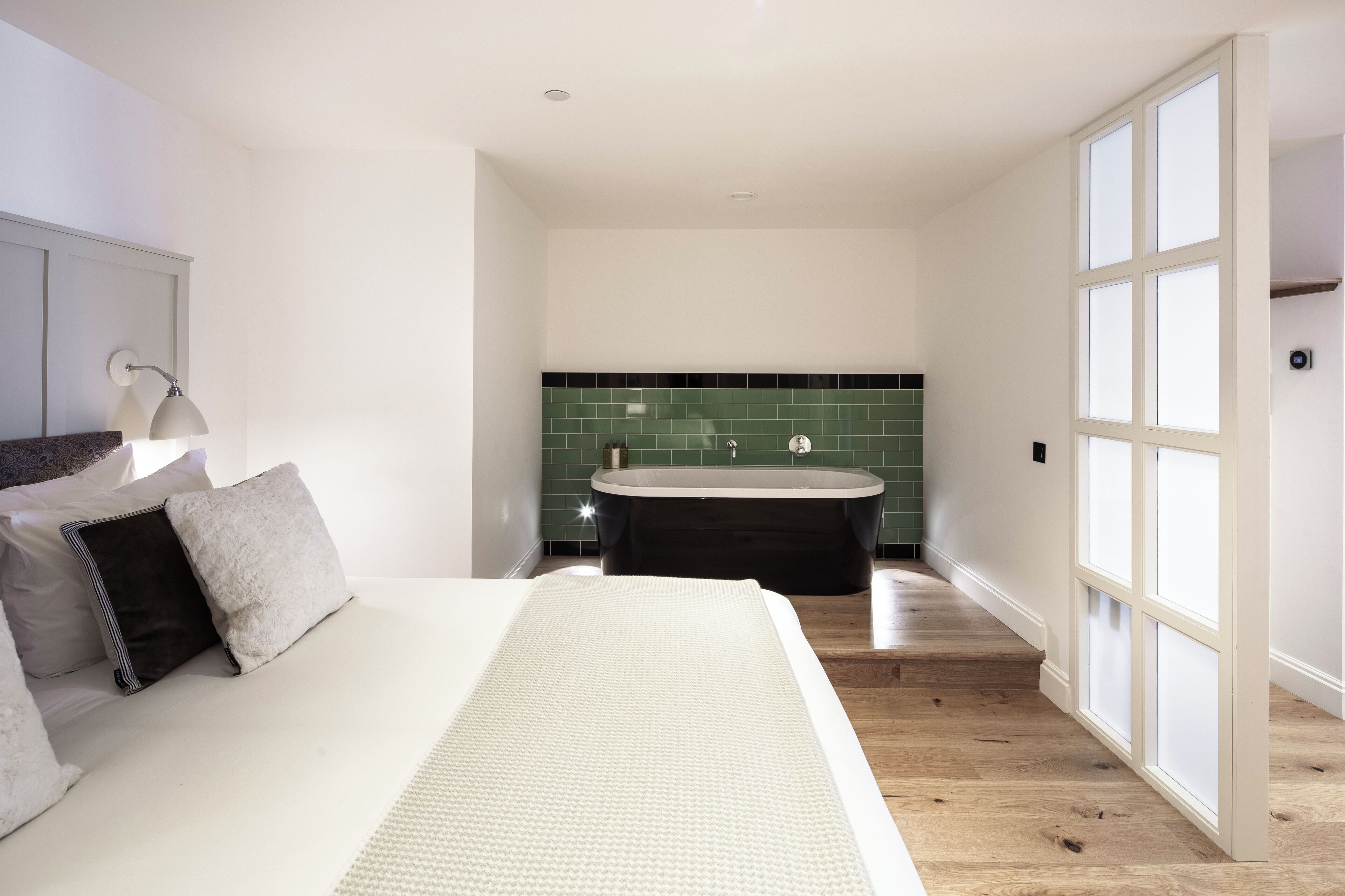 A modern bedroom with a large bed, a nightstand, a lamp, and a view of a bathroom with a bathtub and green tiles.