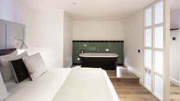 A modern bedroom with a large bed, a nightstand, a lamp, and a view of a bathroom with a bathtub and green tiles.