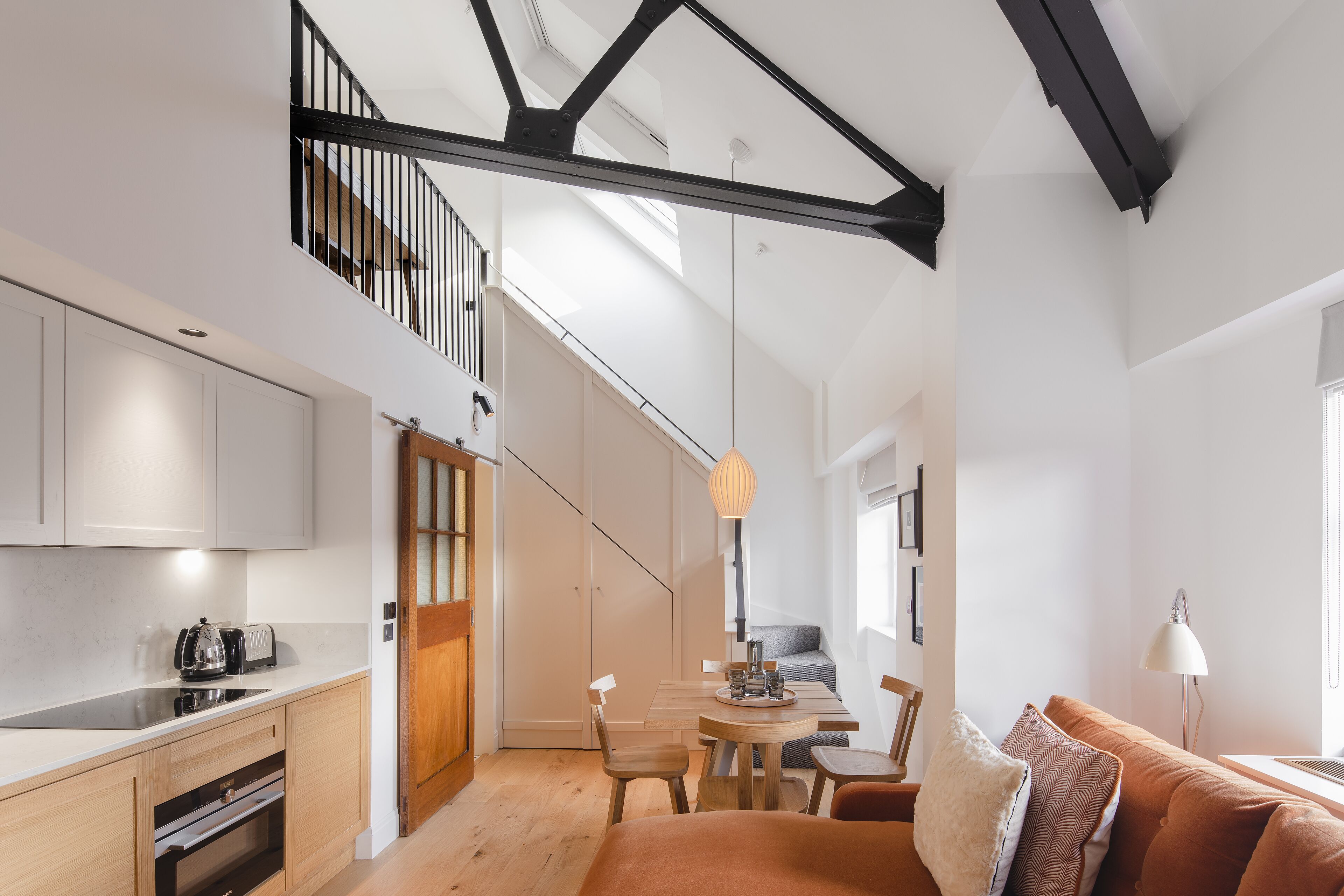 A modern kitchen with a built-in oven, a dining area with a wooden table and chairs, and a loft bedroom with a skylight.