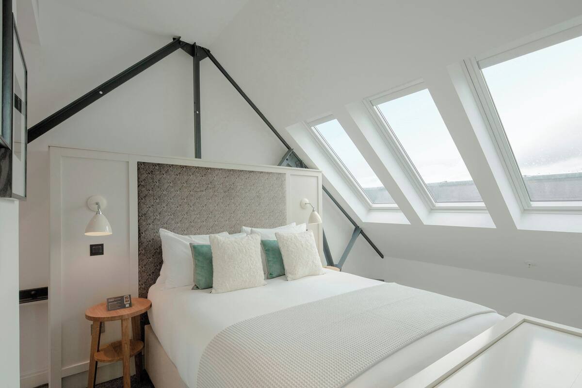A modern hotel room with a large bed, skylights, a headboard with a textured design, a bedside table with a lamp, and a small radio.