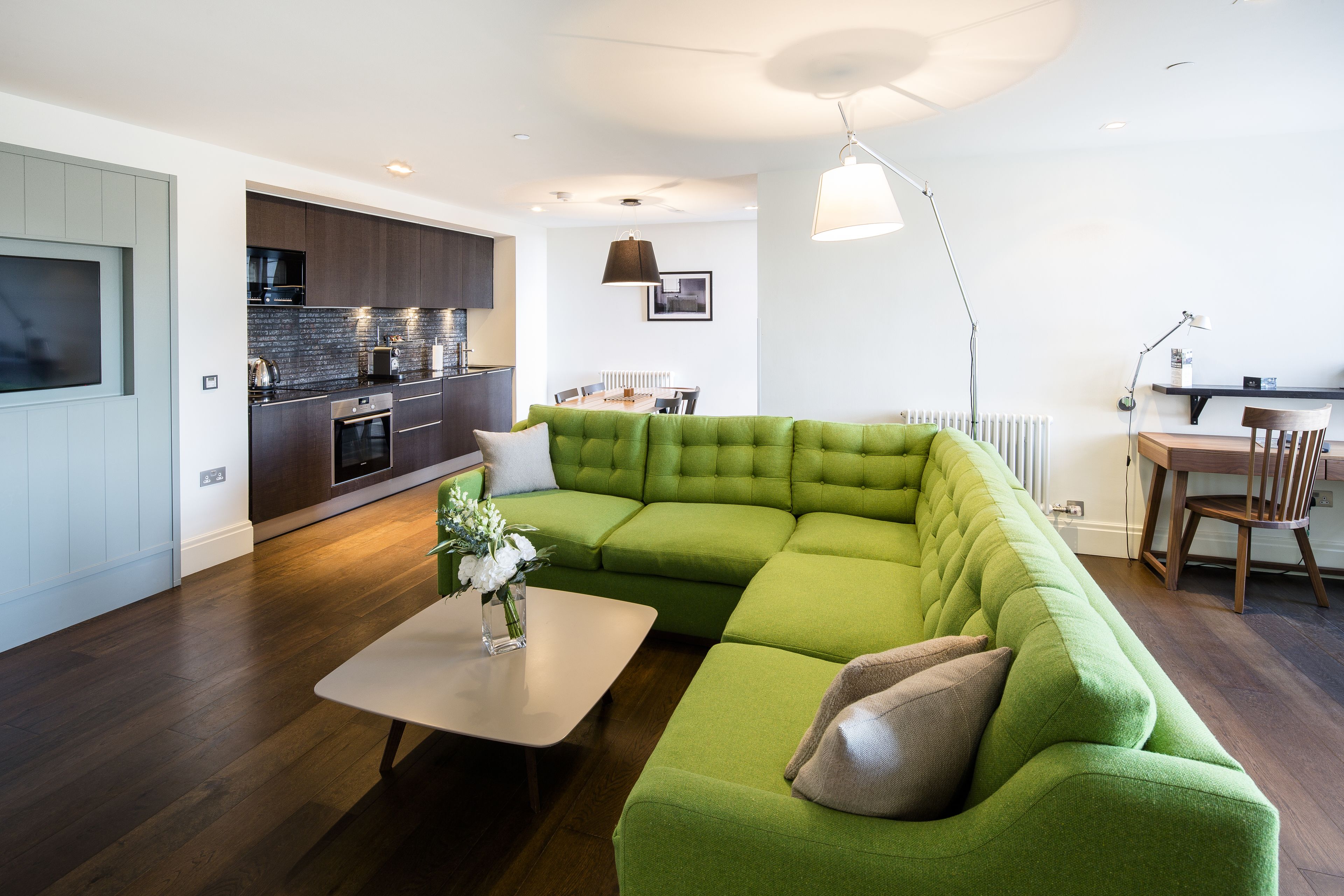 Uma sala de estar moderna com um sofá seccional verde, uma mesa de jantar e uma área de cozinha com armários escuros e bancada branca.