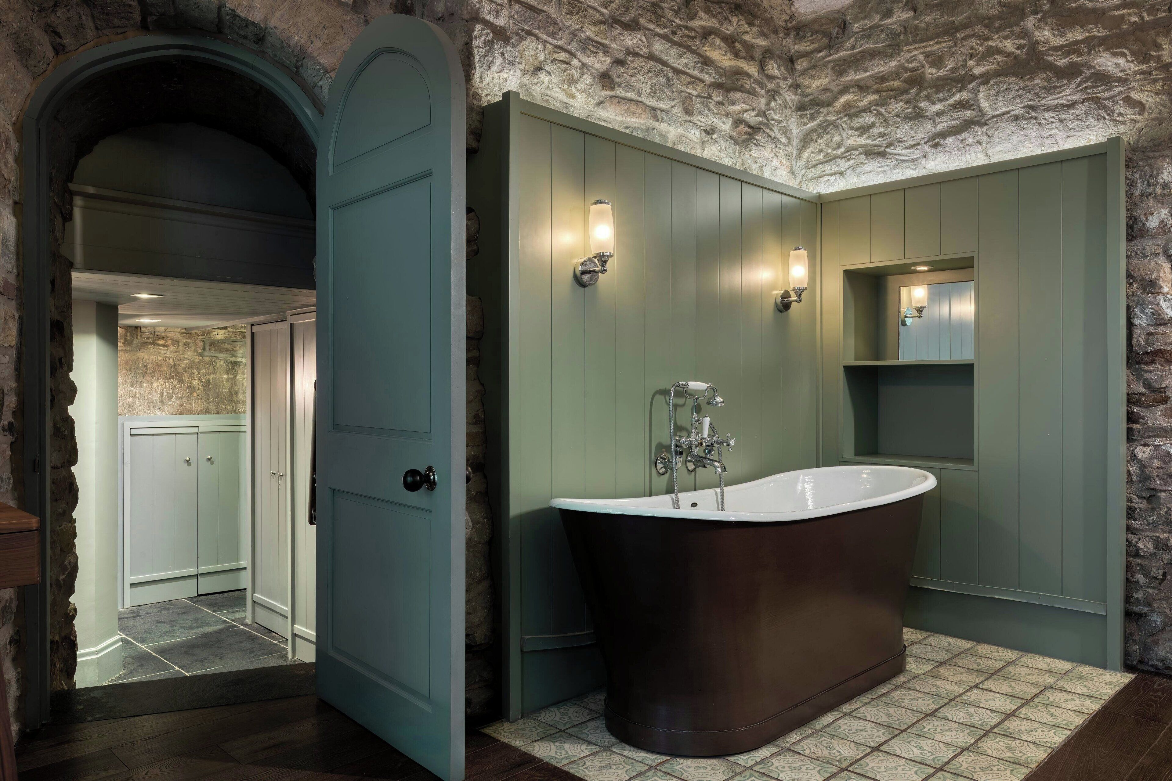 A bathroom with a freestanding bathtub, a stone wall, and a door leading to another room.
