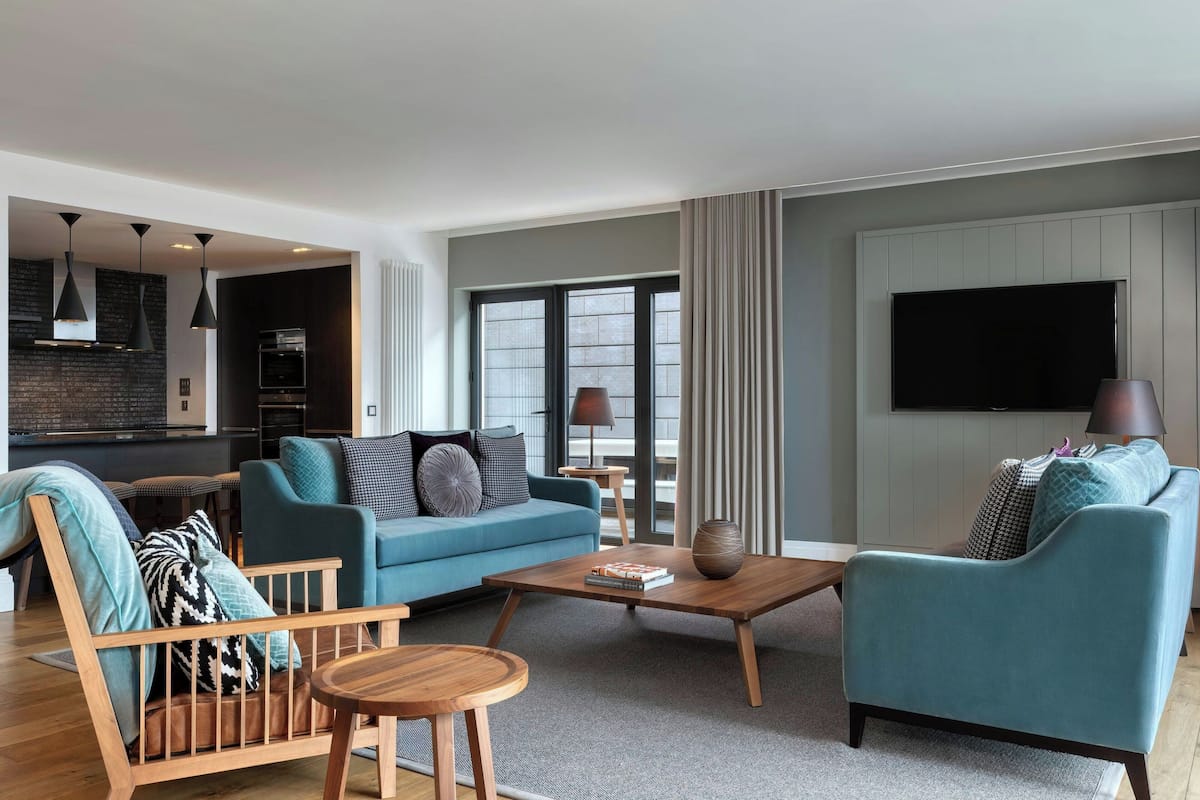 A modern living room with a blue sofa, a wooden coffee table, and a wooden chair. There is a television mounted on the wall and a kitchen area in the background.