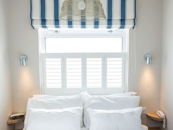 Un dormitorio con cama, una ventana con contraventanas, una lámpara colgante y mesitas de noche con teléfono y lámpara.