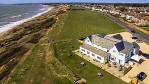 An aerial view of a coastal property with a large grassy area, a white building with multiple rooms, and a road running alongside the beach. - The Cliff House Hotel (New Milton)