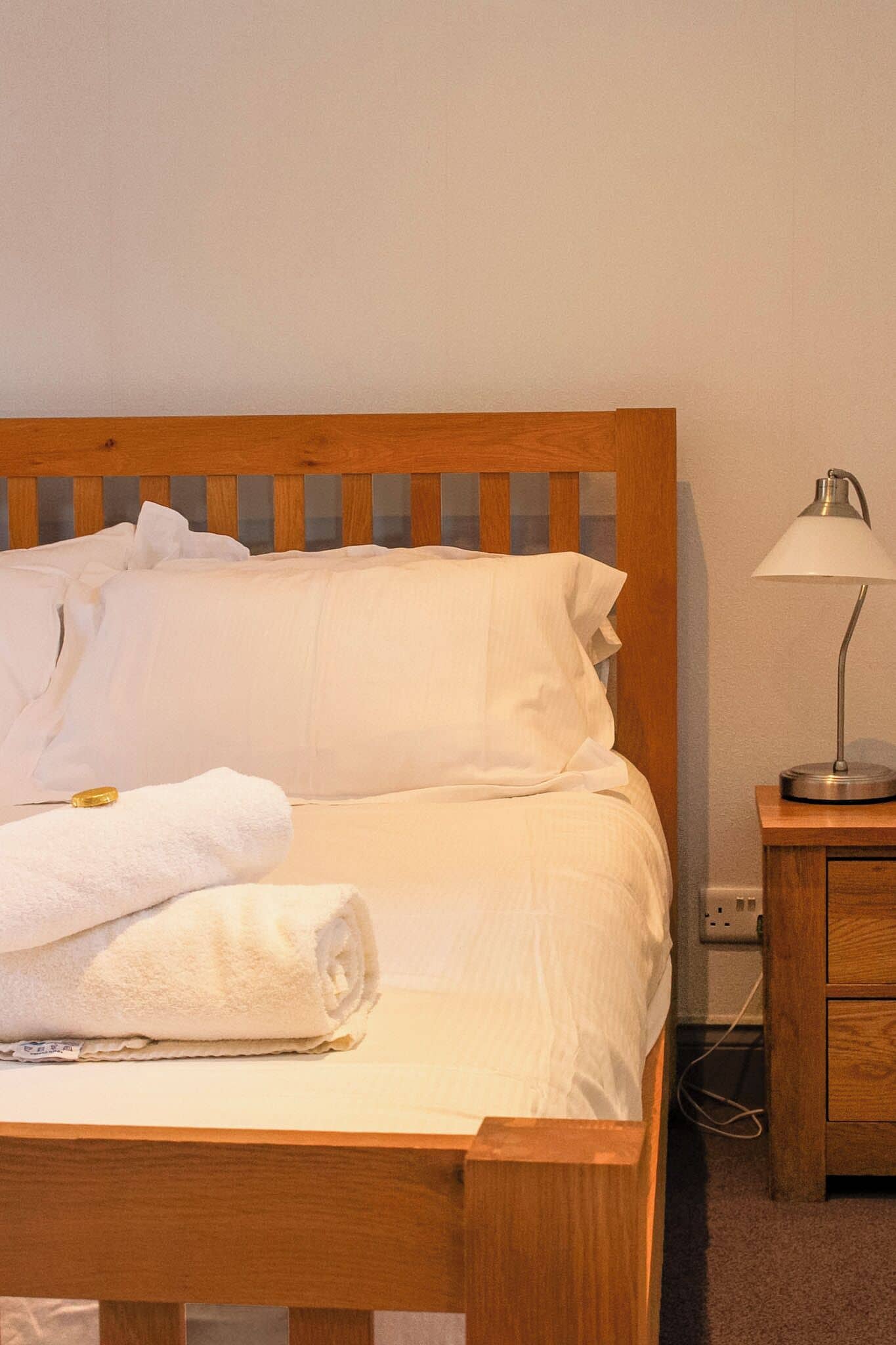 Een netjes opgemaakt bed met witte lakens en een opgerolde handdoek, een houten hoofdbord en een nachtkastje met een lamp.