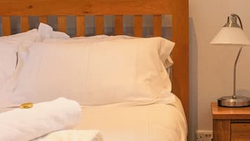 Een netjes opgemaakt bed met witte lakens en een opgerolde handdoek, een houten hoofdbord en een nachtkastje met een lamp.