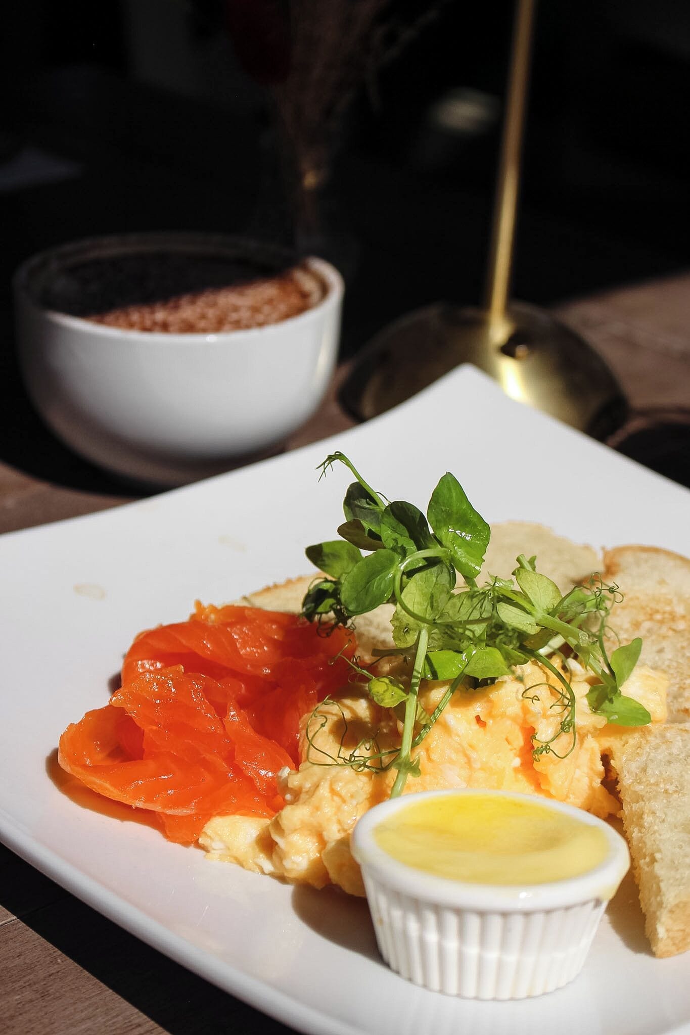Un plato de comida con salmón, huevos revueltos y verduras, acompañado de un recipiente pequeño de salsa y una taza de café.