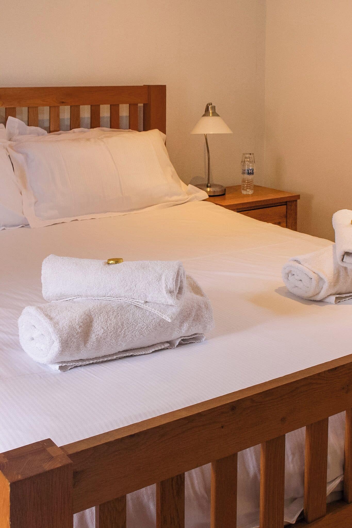 Una cama bien hecha con sábanas blancas, cabecera de madera, mesita de noche con lámpara y una botella de agua.