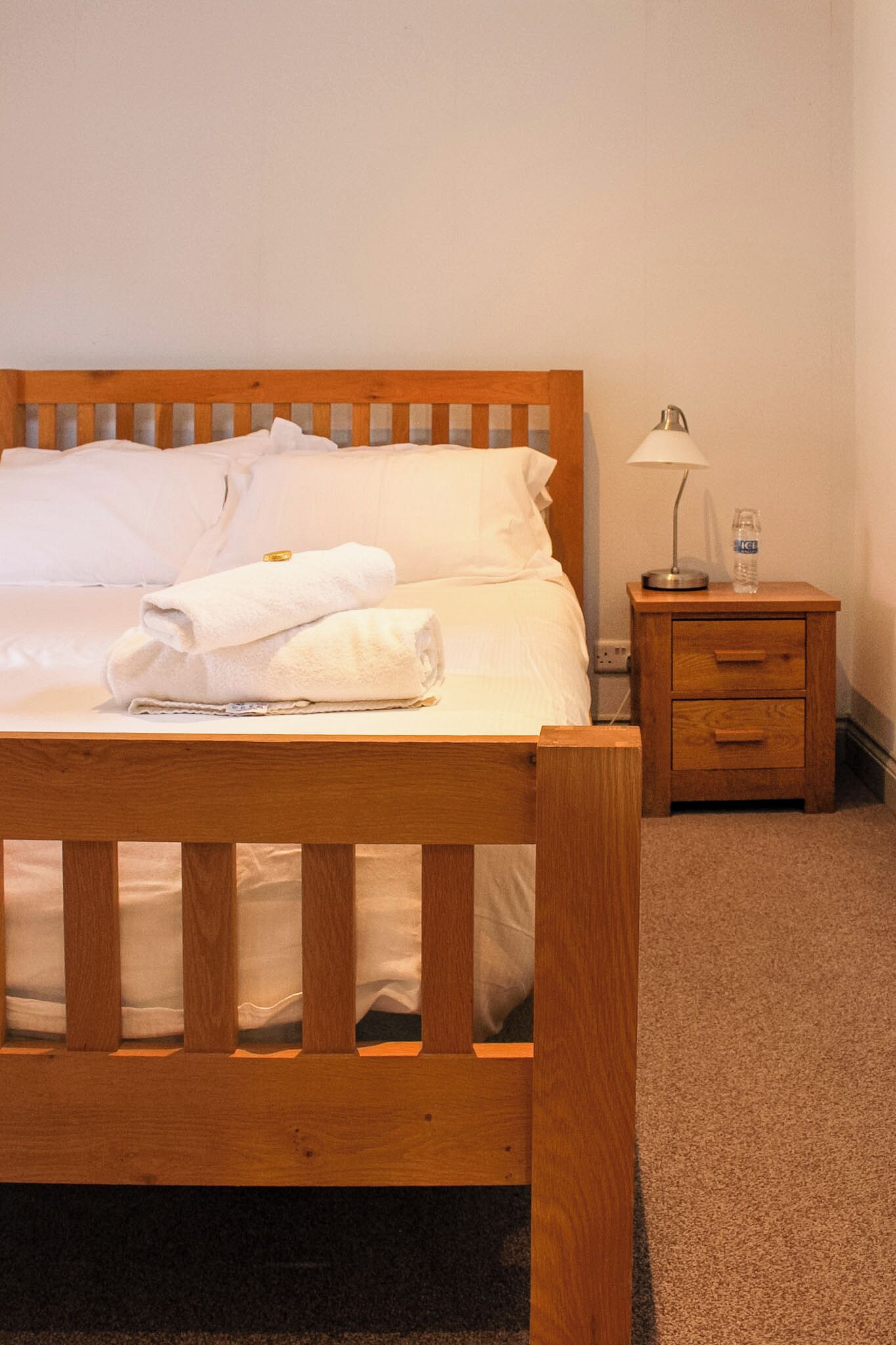 Un lit bien fait, avec du linge de lit blanc et une serviette pliée, une table de chevet avec une lampe et une bouteille, et un sol recouvert d’une moquette.