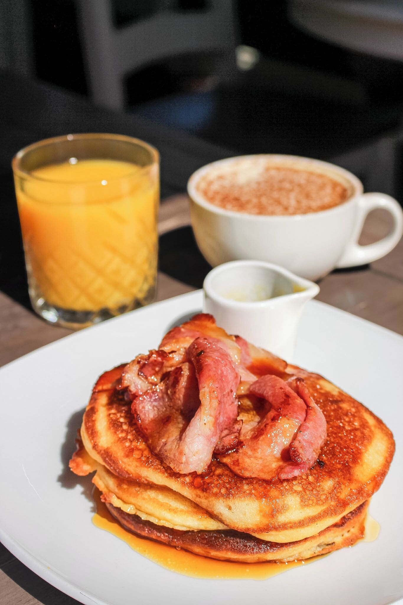 Un plato de panqueques con tocino y jarabe, un vaso de jugo de naranja y una taza de café con azúcar.