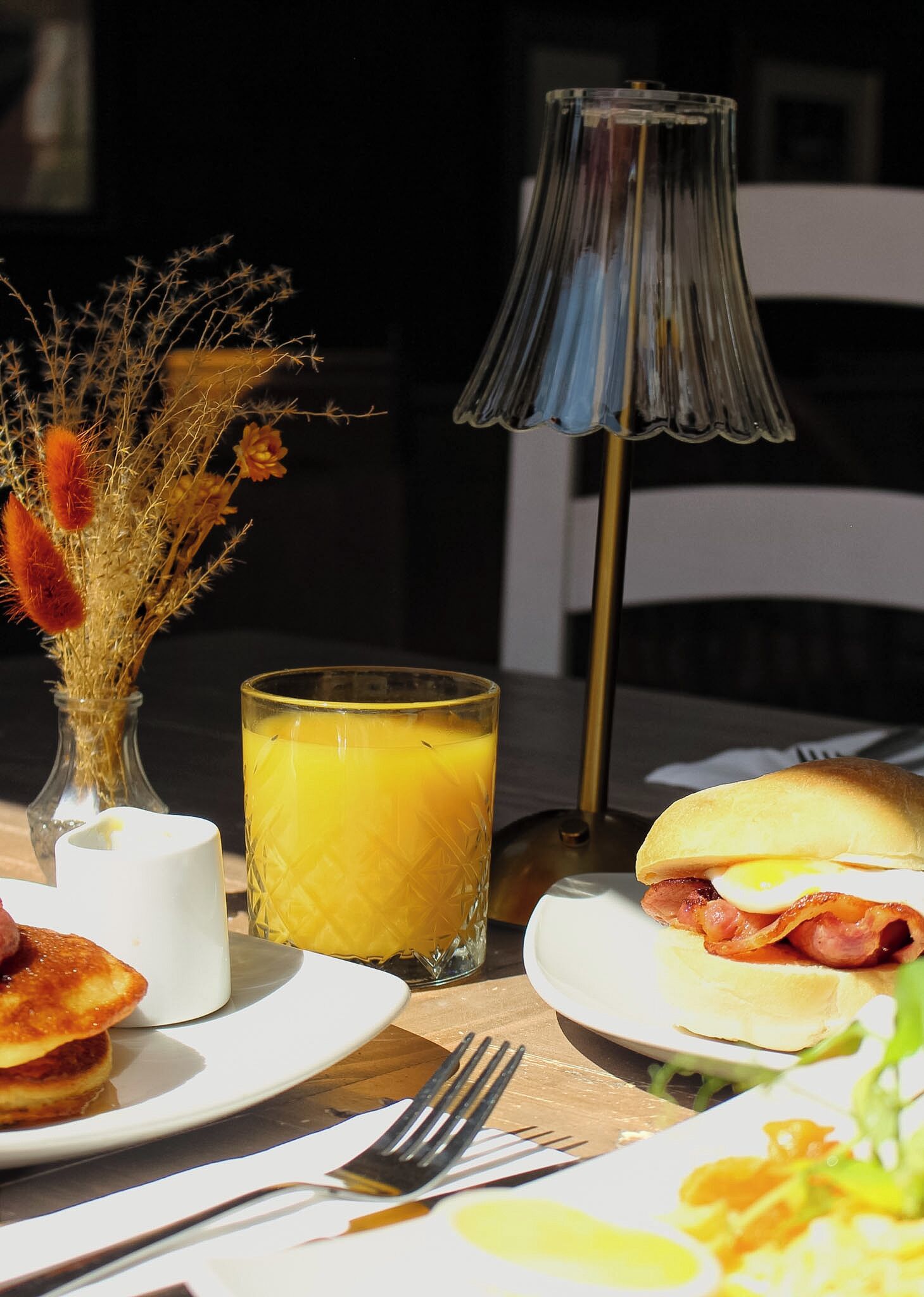 A breakfast setting with a glass of orange juice, a plate with a sandwich, and a vase with dried flowers.