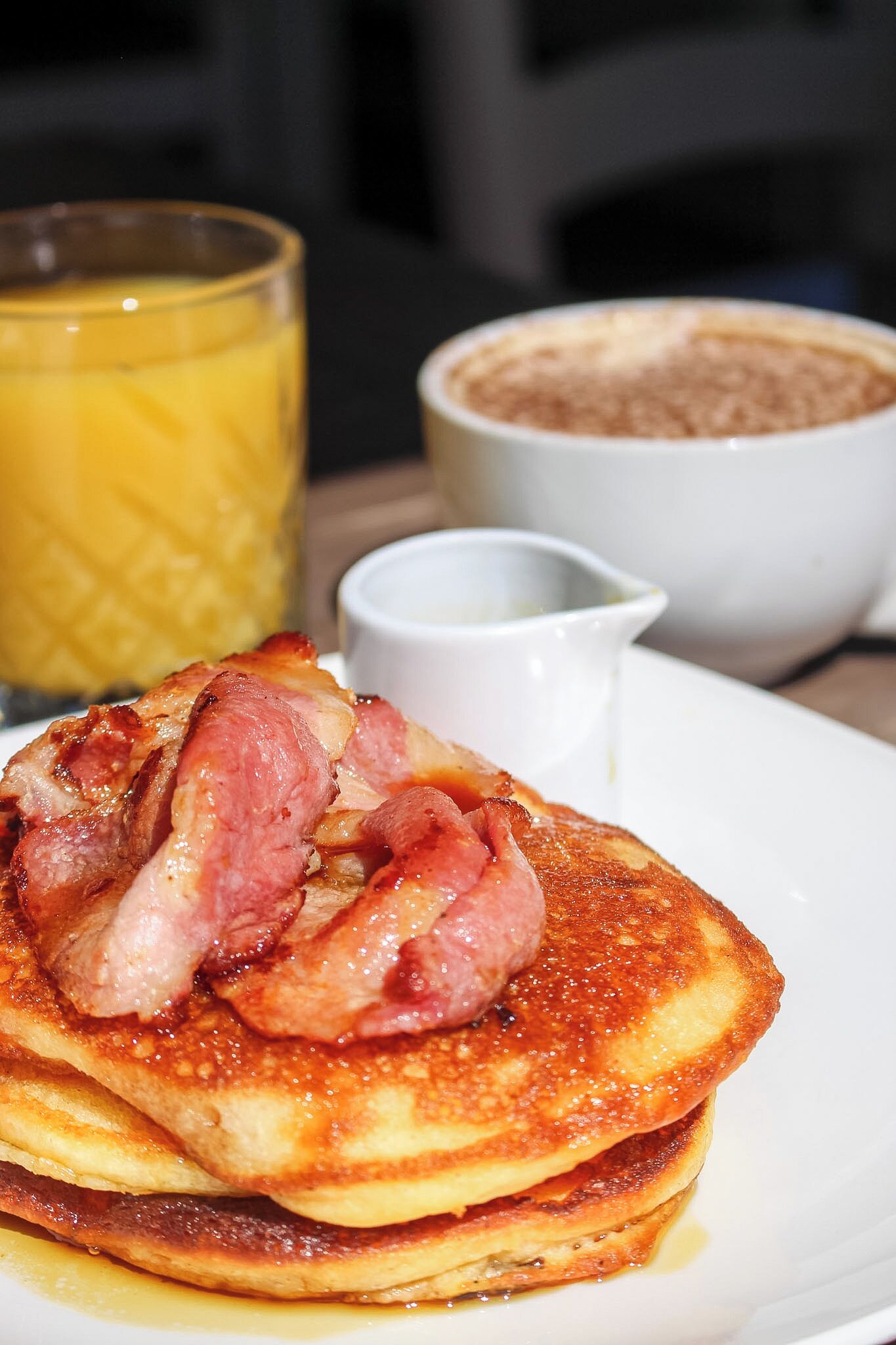 A plate of pancakes with bacon, a glass of orange juice, and a bowl of what appears to be cinnamon sugar.