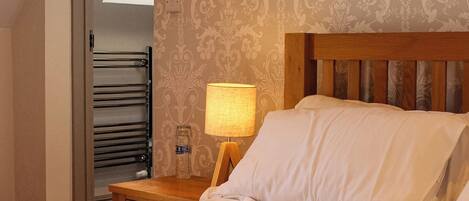 A neatly made bed with white linens and a rolled towel, a wooden headboard, a nightstand with a lamp, and a patterned wallpaper.