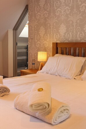 A neatly made bed with white linens and a rolled towel, a wooden headboard, a nightstand with a lamp, and a patterned wallpaper. - The Selsey Arms (Chichester)