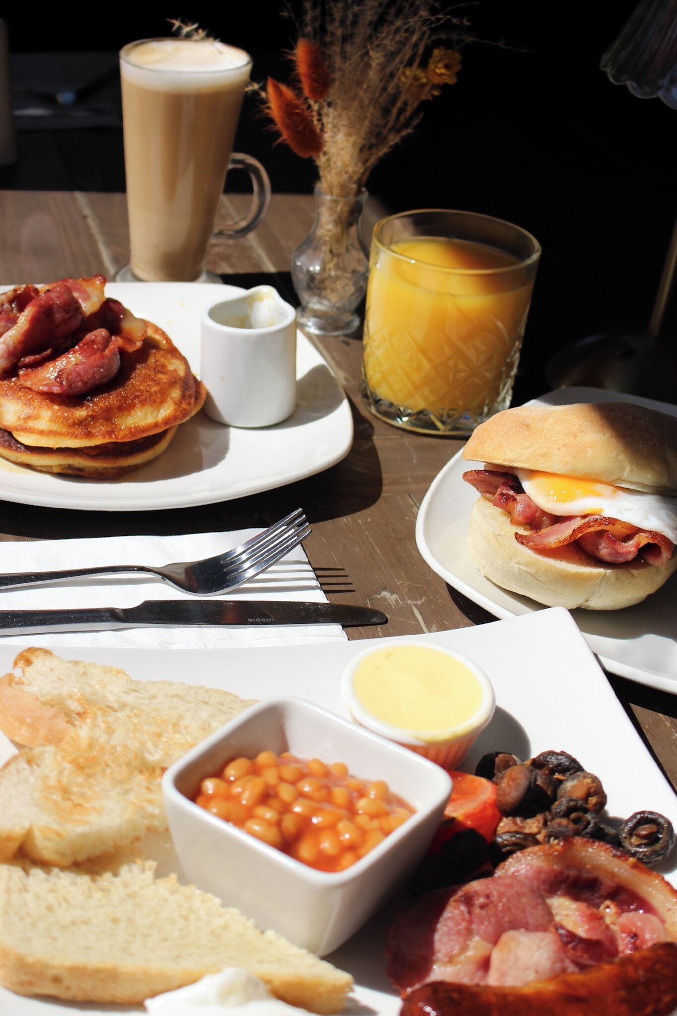 A breakfast table with pancakes, eggs, bacon, toast, beans, and a glass of orange juice.