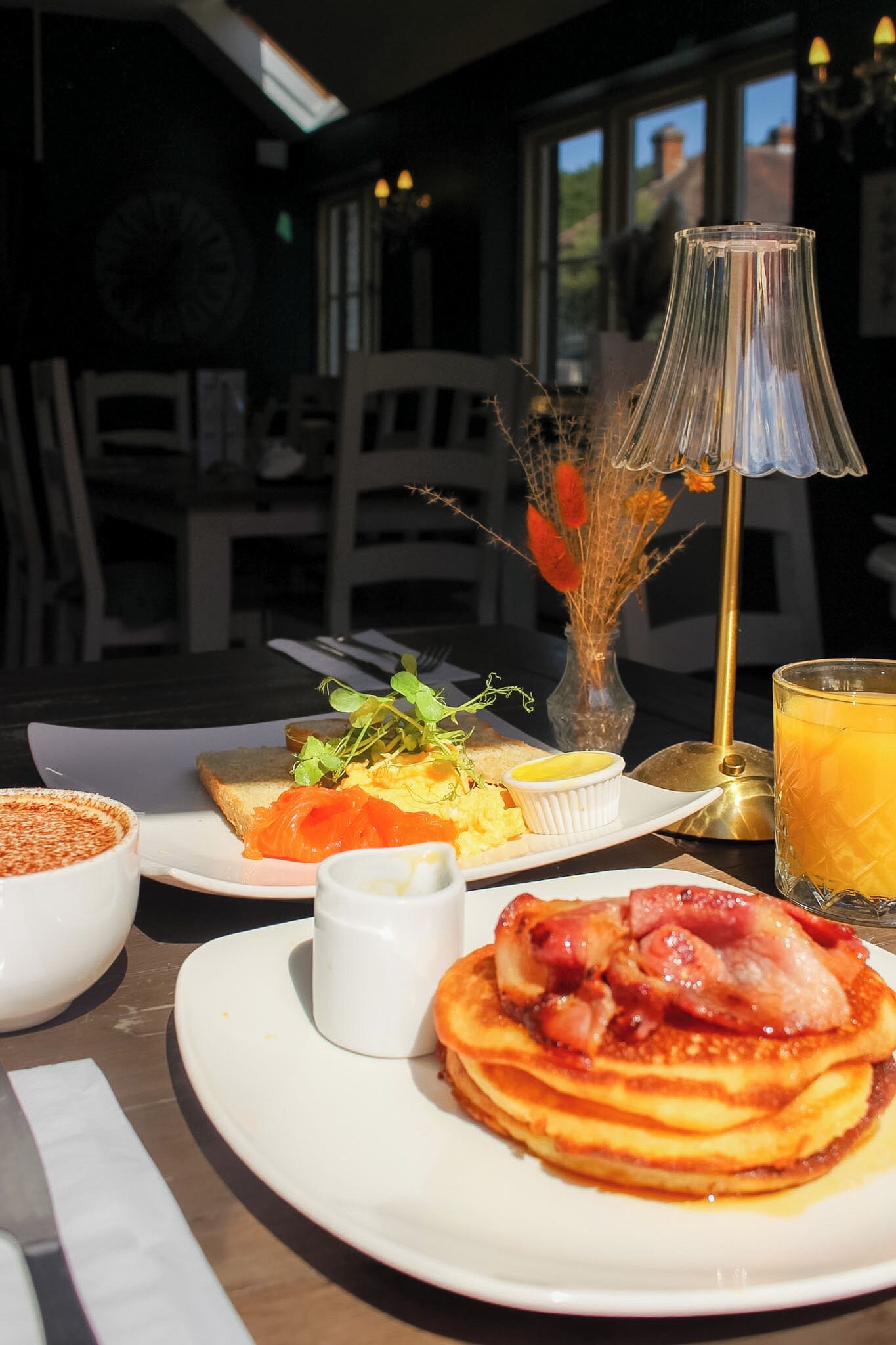 A breakfast setting with a plate of pancakes, a glass of orange juice, and a small vase with dried flowers.