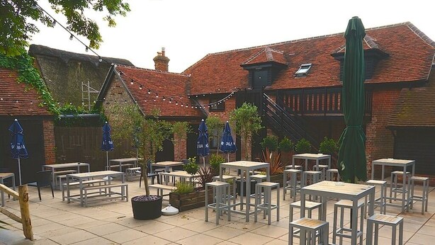 An outdoor patio with multiple tables and chairs, surrounded by brick buildings with a traditional design. - The Selsey Arms (Chichester)