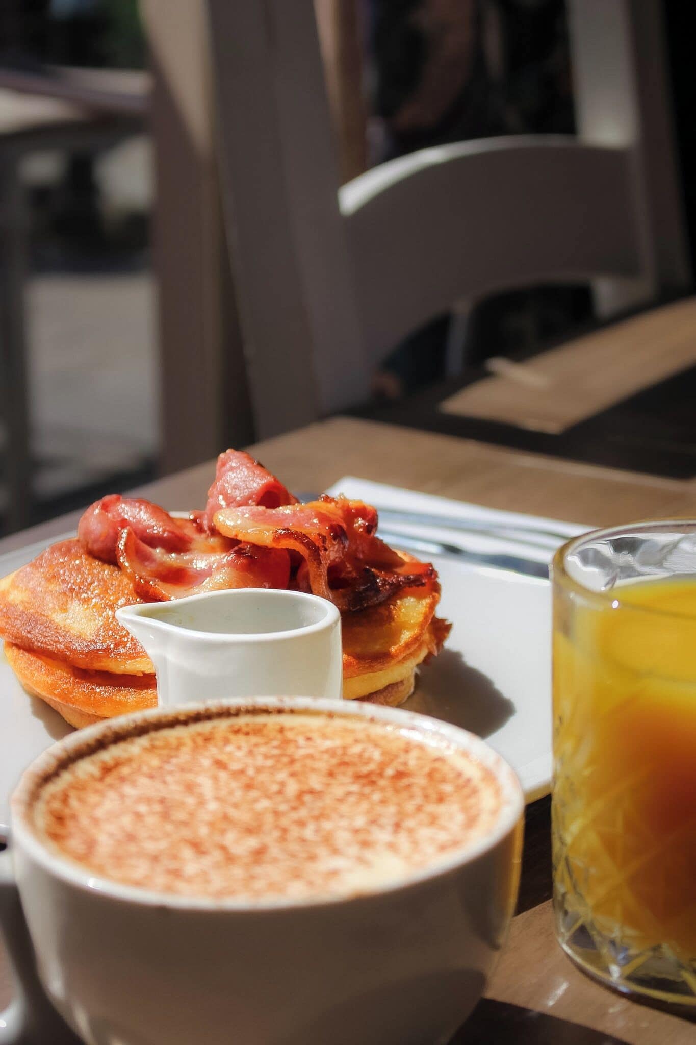 Une table dressée pour le petit-déjeuner avec des crêpes, du bacon, une tasse de café et un verre de jus d’orange.