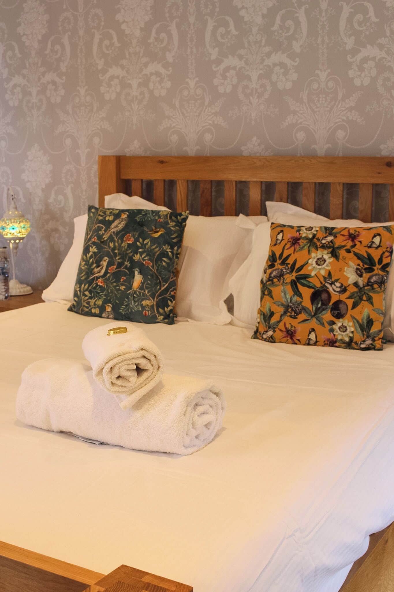 Ein ordentlich bezogenes Bett mit weißen Leinen, zwei dekorative Kissen mit floralem Muster und ein aufgerolltes weißes Handtuch.