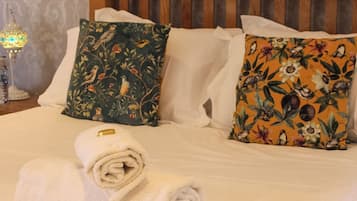 Ein ordentlich bezogenes Bett mit weißen Leinen, zwei dekorative Kissen mit floralem Muster und ein aufgerolltes weißes Handtuch.