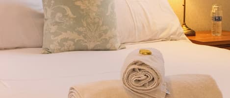 Una cama bien hecha con una toalla enrollada y una almohada, una lámpara de noche y una botella sobre una mesita de noche.