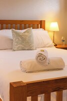 Una cama bien hecha con una toalla enrollada y una almohada, una lámpara de noche y una botella sobre una mesita de noche.