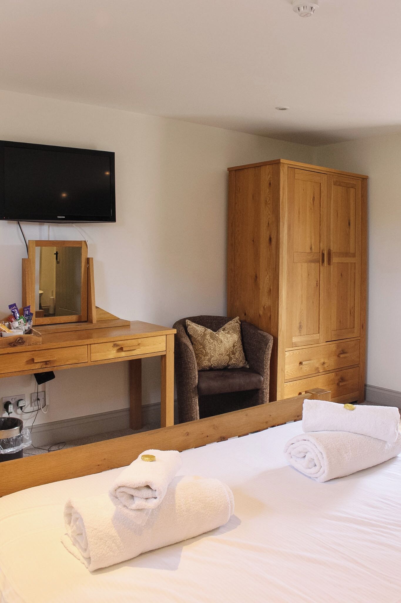 A hotel room with a bed, a wooden wardrobe, a desk with a mirror, and a television.