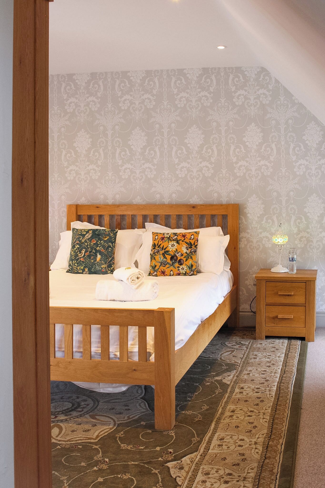 A bedroom with a wooden bed, patterned pillows, a bedside table with a lamp, and a decorative rug.