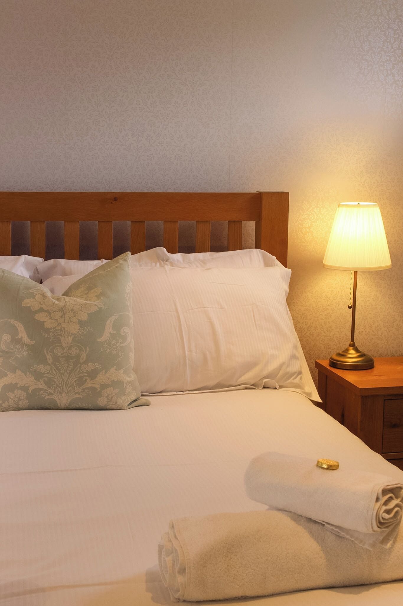 Una cama bien hecha con sábanas blancas, una almohada con estampado floral verde y una toalla doblada al lado.