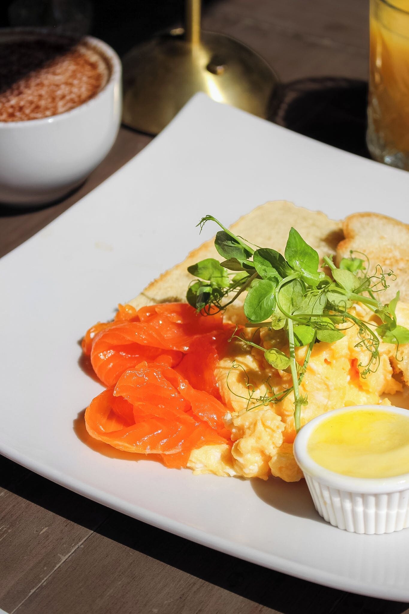 A plate with scrambled eggs, smoked salmon, and a small bowl of sauce, served on a white plate.
