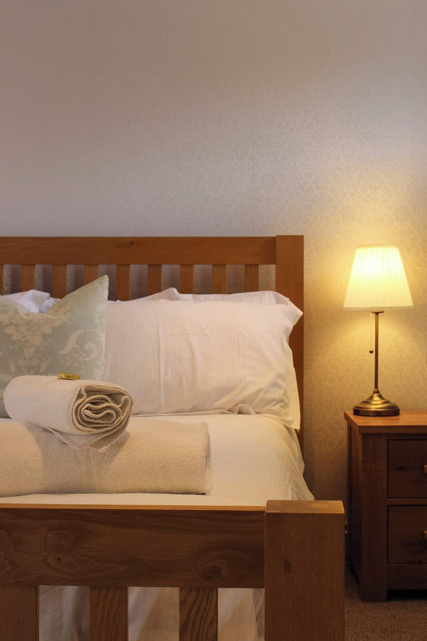 A neatly made bed with a rolled towel, a bedside lamp, and a wooden nightstand.