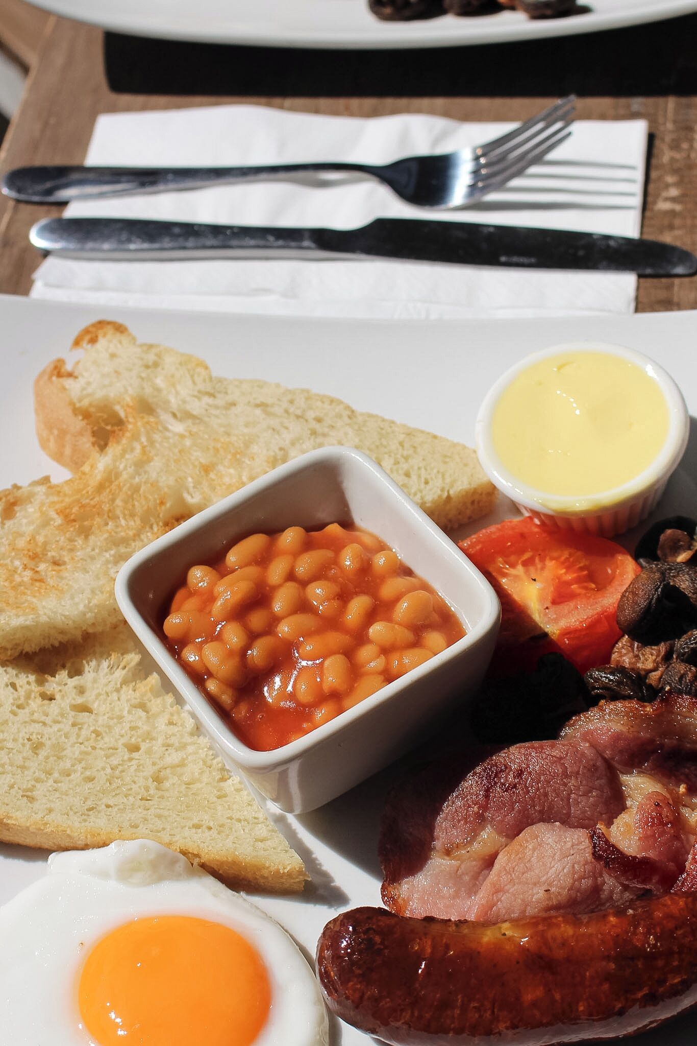 A breakfast plate with toast, beans, bacon, sausages, a fried egg, and a small bowl of butter.