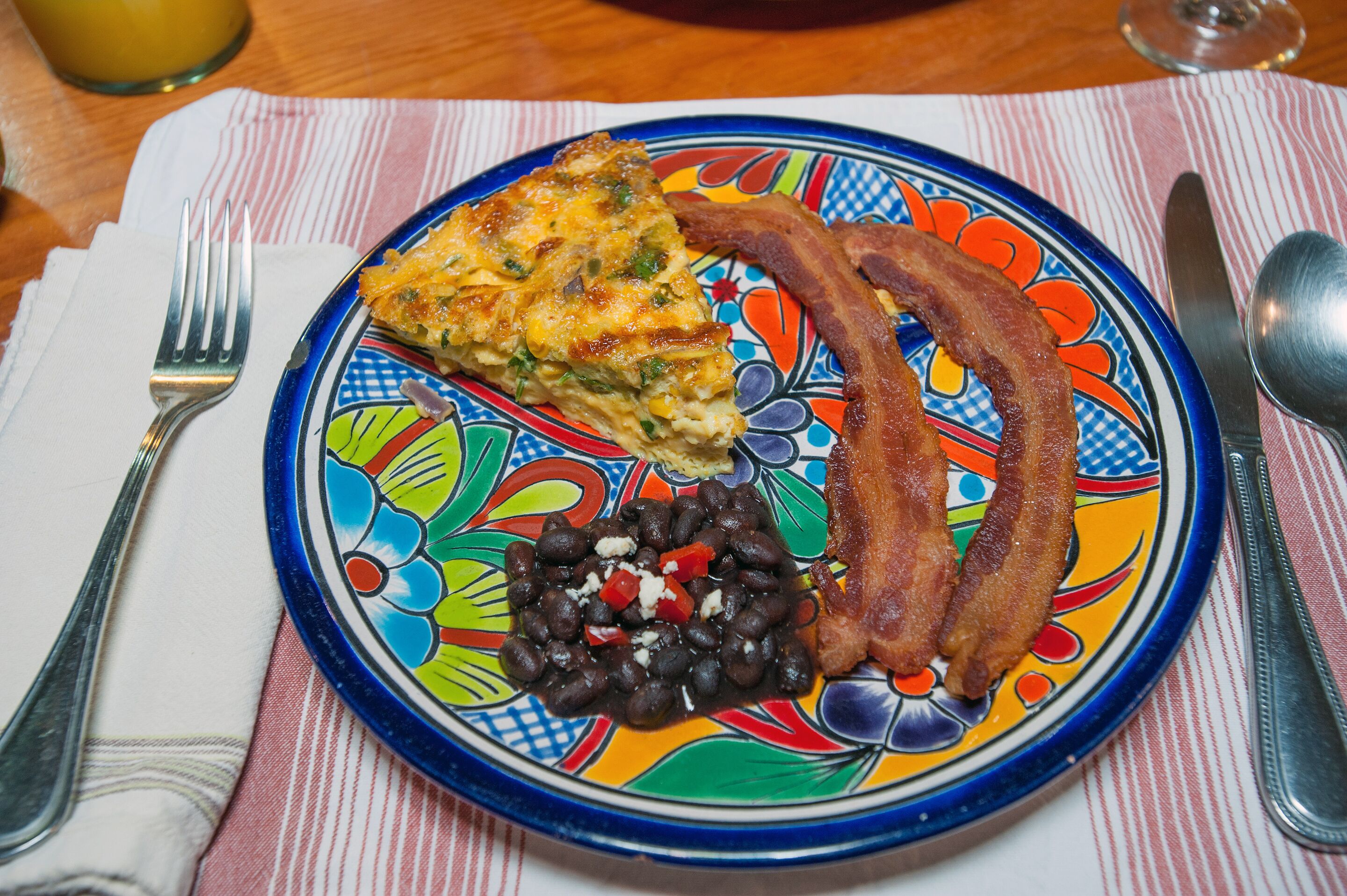 Un plato con una porción de quiche, dos tiras de tocino y una guarnición de frijoles negros con pimientos rojos.