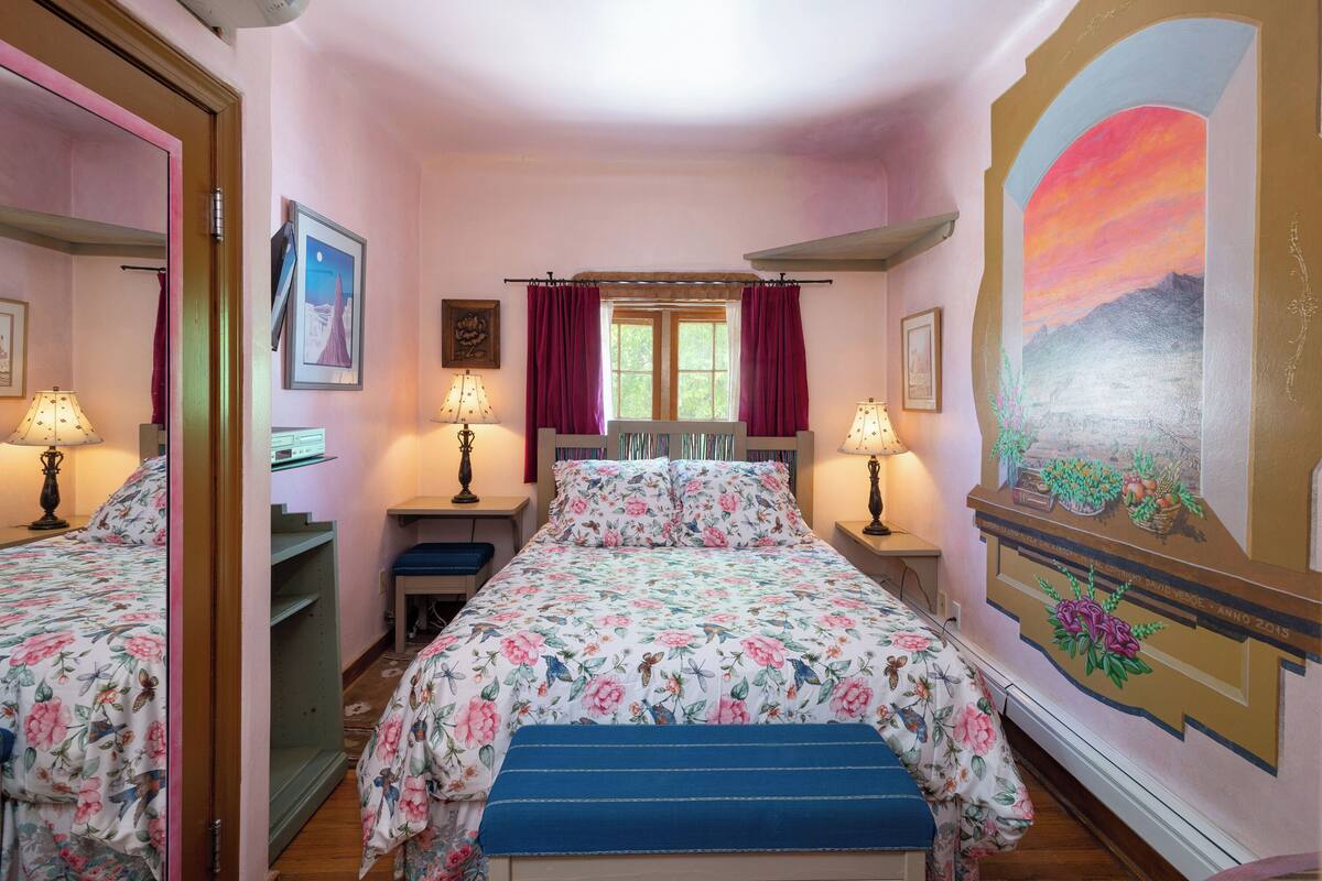 A bedroom with a floral bedspread, a bench, a window with curtains, and a mural on the wall.