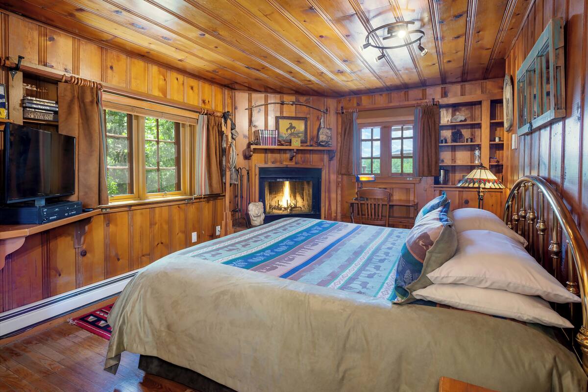 A wooden cabin bedroom with a bed, a fireplace, a television, and a window with curtains.