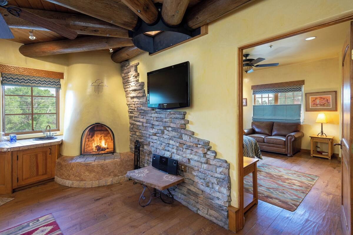A cozy living room with a stone fireplace, wooden ceiling beams, a flat-screen TV mounted on the wall, and a comfortable seating area with a couch and a small table.