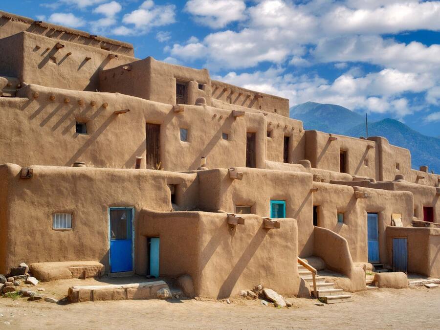A traditional adobe building with multiple doorways and windows, set against a mountainous backdrop.