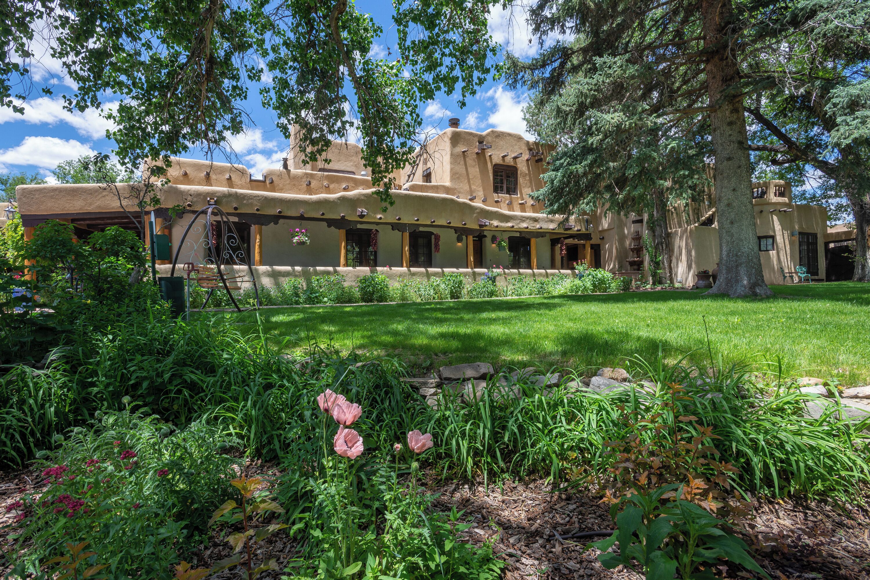 A traditional adobe building with a covered patio area, surrounded by lush greenery and flowering plants.