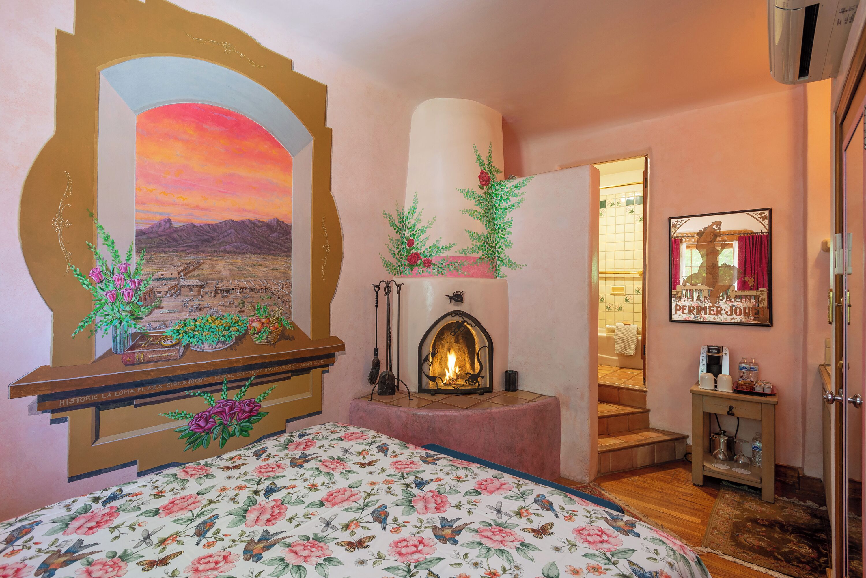 A bedroom with a fireplace, floral bedspread, and a painting of a sunset.