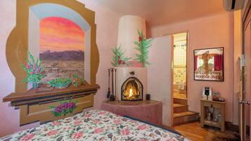 A bedroom with a fireplace, floral bedspread, and a painting of a sunset.